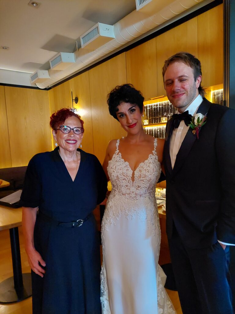 wedding officiant SandraLaya Ruch stands beside bride with short brown hair wearing a traditional wedding dress and groom stands beside her in a black suit, bowtie and a white boutineer.