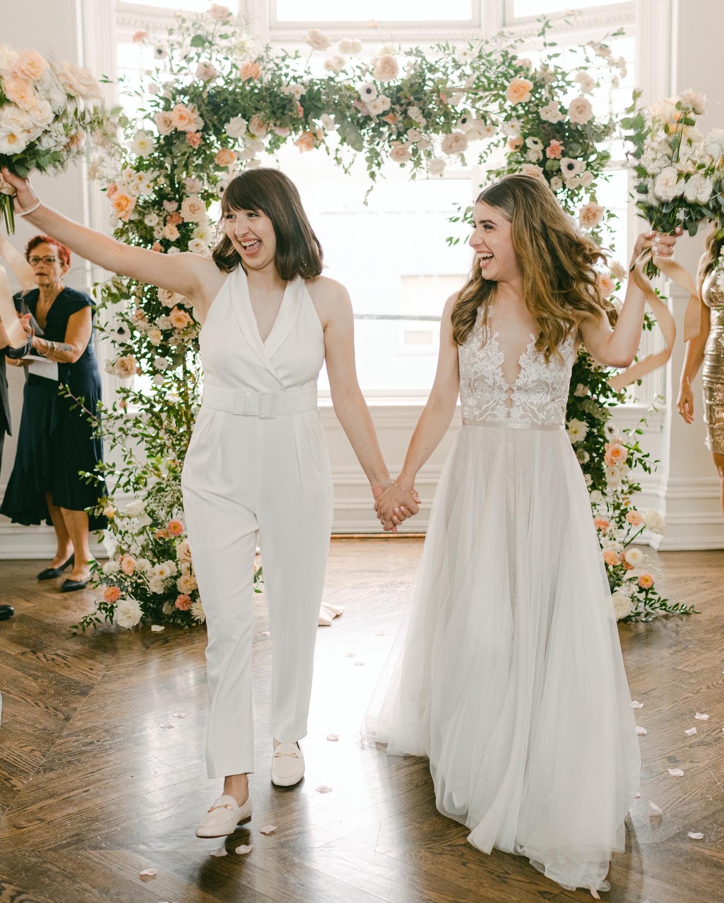 2 beautiful brides both wearing white. 1 bride is wearing a sleeveless jumpsuit and the other bride is wearing a long white dress. Couple is holding hands with the wedding canopy (chupa) in the background.