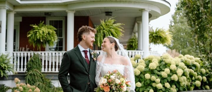 wedding day for Erica and Rykker. Beautiful bride in white wedding dress looks lovingly at her handson groom in a dark coloured suit in front of a beautiful 2 story wedding venue with 2 levels of white verandas.