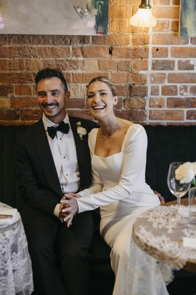 Bride in traditional white wedding dress sitting and holding hands with groom dressed in a black tux with bowtie.