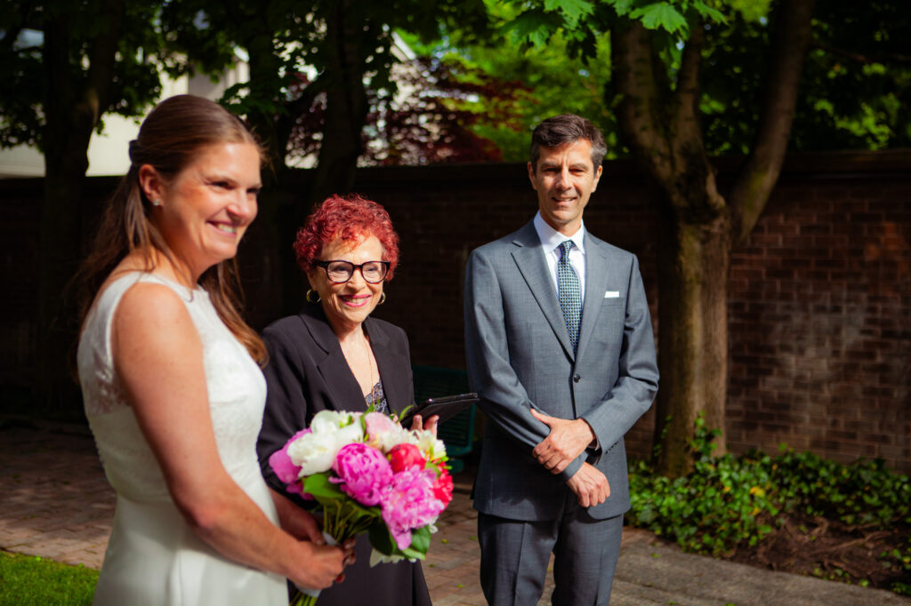 Bride in white wedding dress with bouquet of pink and white flowers standing beside SandraLaya Ruch, wedding and groom dressed in a grey suit and tie.